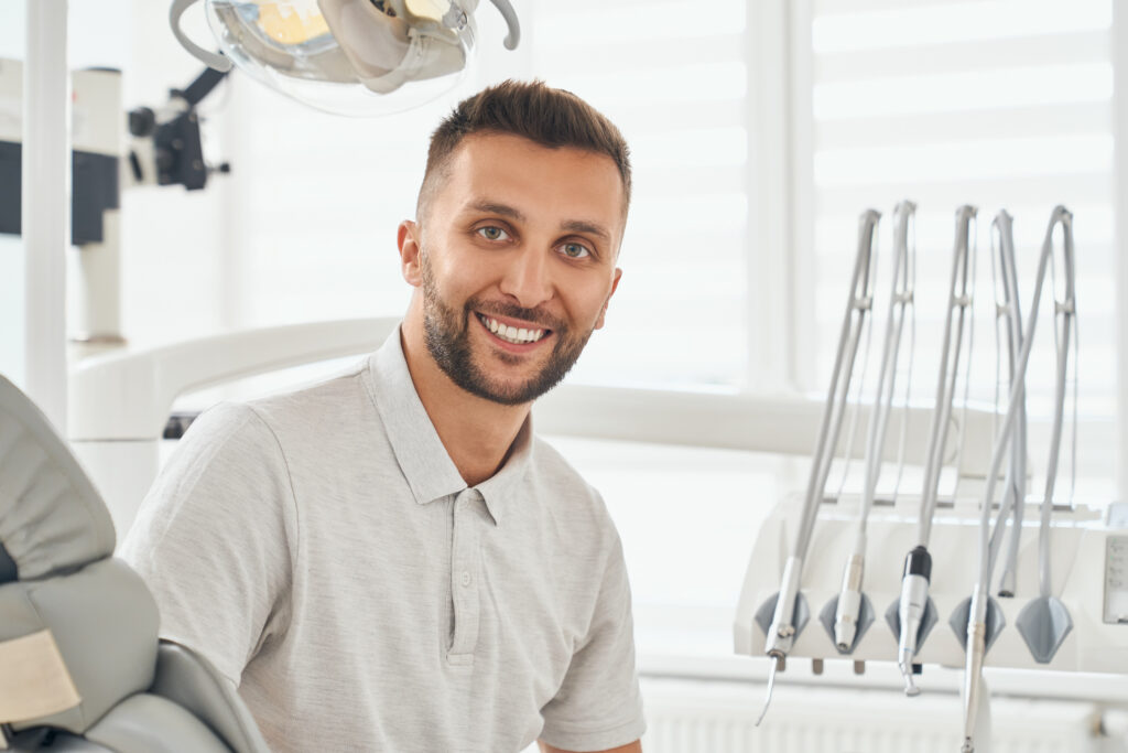 Portrait of happy bearded man in grey t-shirt sitting in dental chair, smiling and looking at camera. Young male patient looking satisfied after hygiene procedure at dental clinic