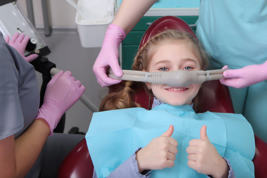 A little girl is comfortable to treat her teeth under superficial sedation. The girl smiles and holds two thumbs up. Milk teeth treatment. Treatment of children's teeth with nitrous oxide. Photos in the interior of the clinic.