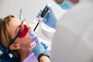 Medic shining with LED curing light on lady teeth for hardening of dental fillings while woman wearing red safety glasses